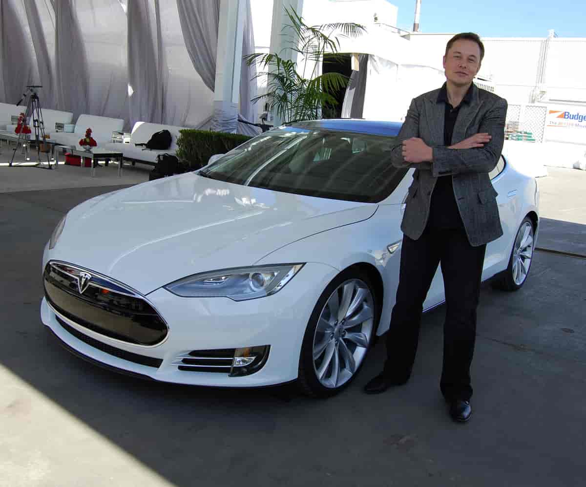 Elon Musk stands confidently in front of a sleek, silver Tesla electric car. He wears a black jacket and holds his hands clasped in front of him, with a small smile on his face. The car is shiny and modern, with clean lines and a futuristic feel.